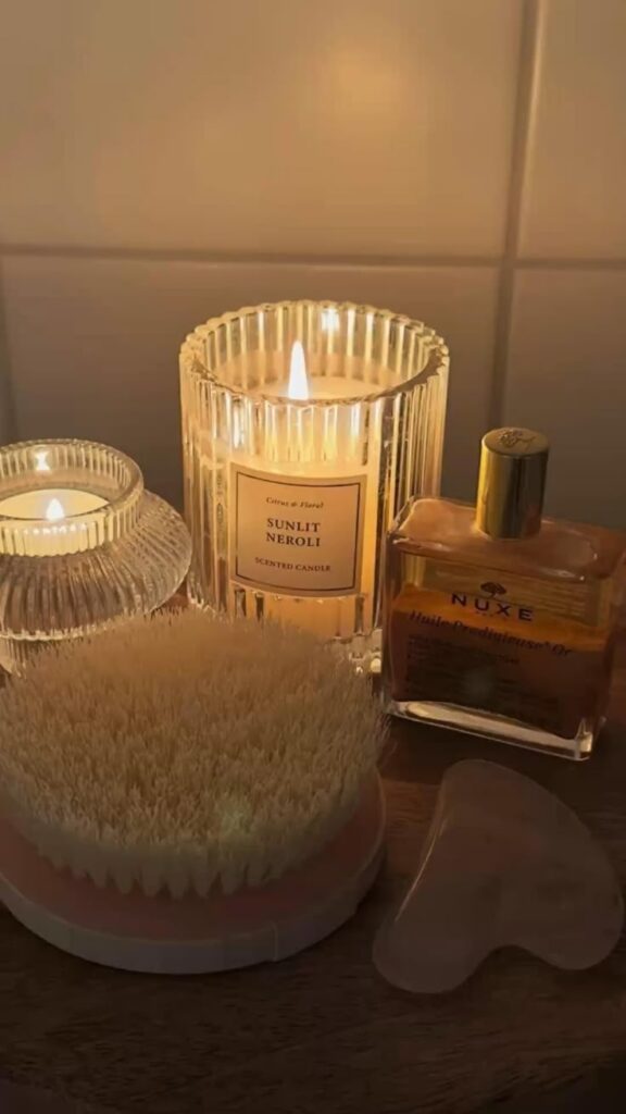 Cozy spa setup with lit candles, dry brush, facial oil, and gua sha stone on a wooden surface for a relaxing self-care ritual.