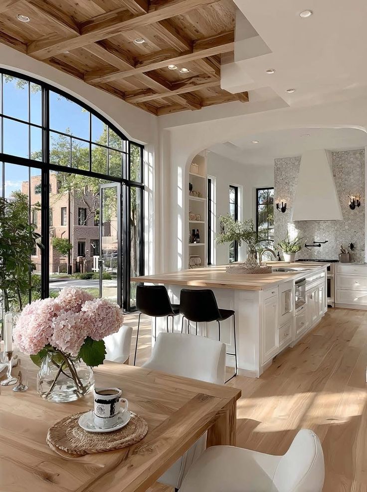 Spacious modern kitchen with a wooden ceiling, large windows, and stylish decor. Bright, airy, with plants and pink flowers on the table.