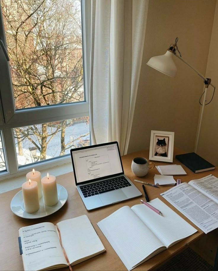 Cozy study desk with laptop, notebook, candles, and window view, ideal for productive work or study sessions.