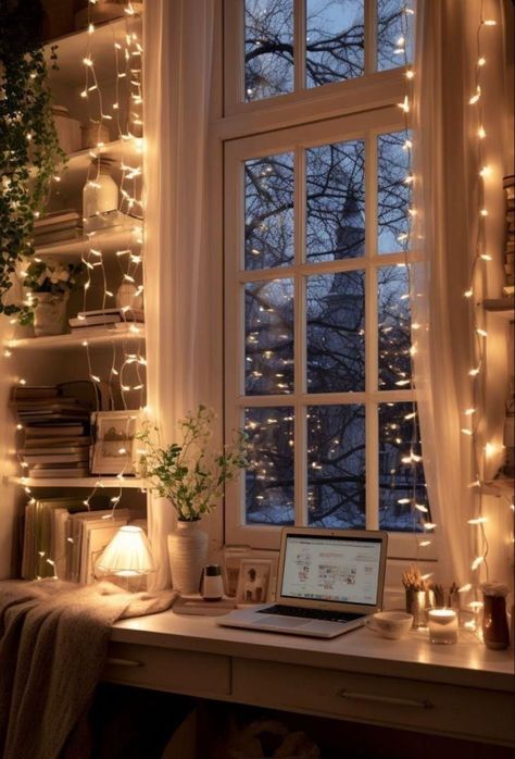 Cozy home office with laptop on desk, surrounded by books, plants, and warm string lights by a large window at twilight.