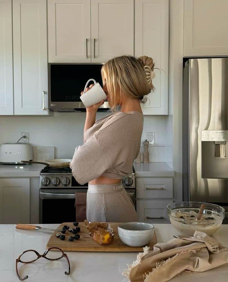 Woman enjoys coffee in cozy kitchen, bowl and fruit on counter. High-key natural lighting, modern appliances.