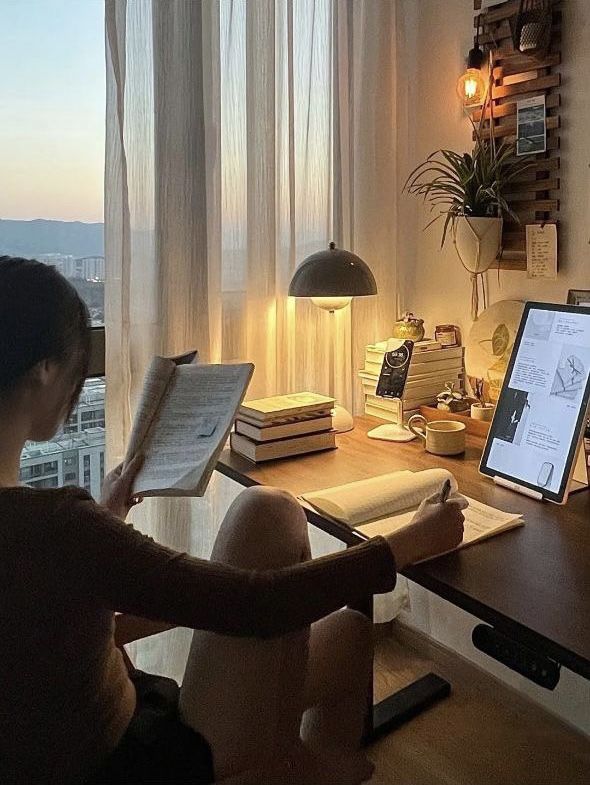 Cozy study setup with a person reading by the window, surrounded by books, a tablet, and warm lighting. Urban sunset view outside.