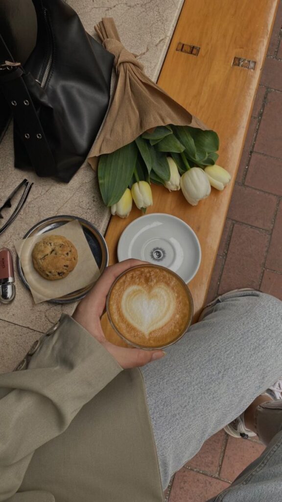 Cozy café moment with heart-shaped latte art, tulips, scone, and bag on a wooden bench. Relax and enjoy the peaceful vibe.
