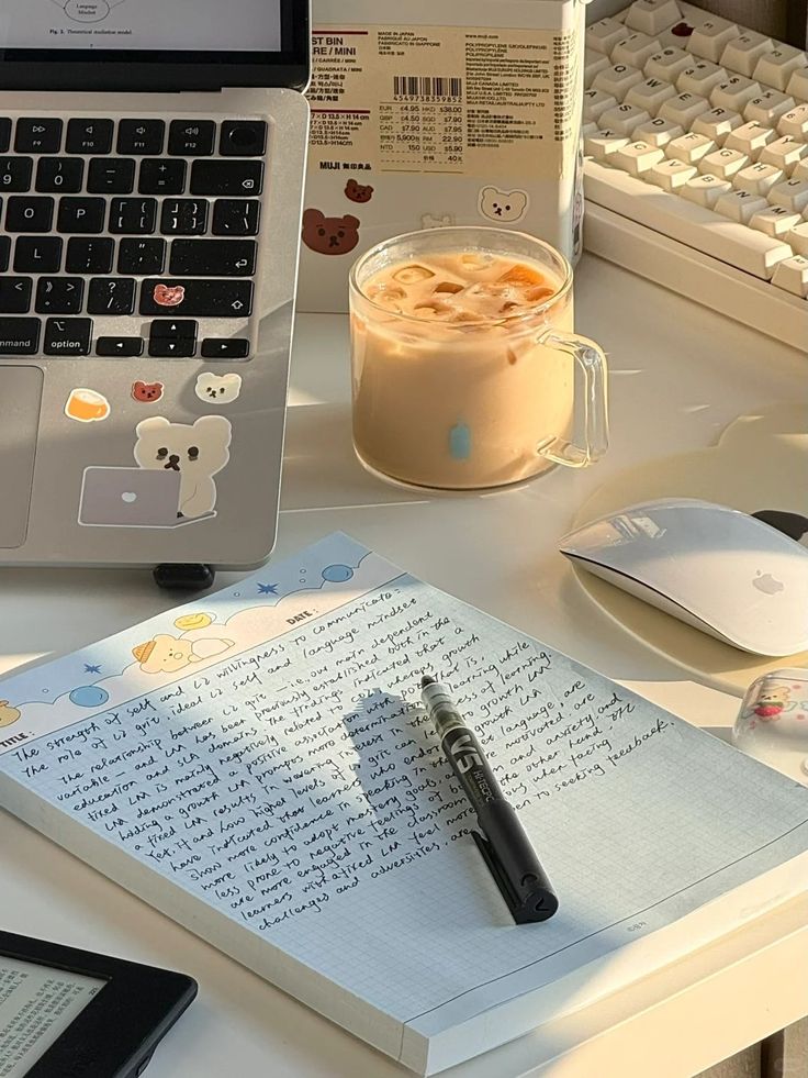 Cozy workstation with a laptop, iced coffee, notebook, and pen on a sunlit desk; perfect for productivity and writing.