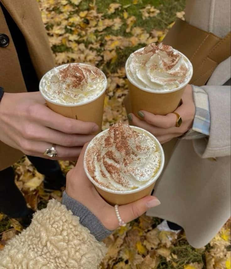Three people holding paper cups of whipped cream-topped lattes outdoors in autumn, surrounded by fallen leaves.