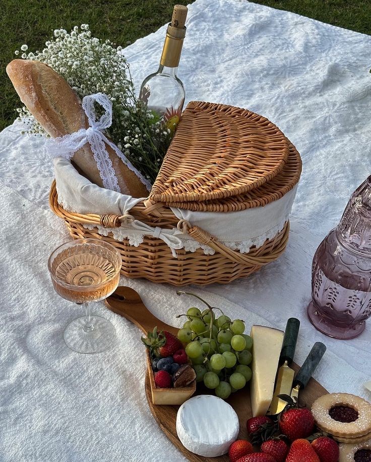 Picnic basket with bread and wine, cheese platter with grapes and berries on a white blanket outdoors. Perfect for a summer picnic.