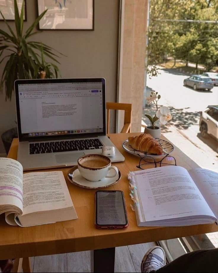 Cozy workspace with a laptop, coffee, books, and a croissant on a wooden table near a sunny window, ideal for productivity.