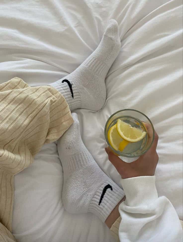 Cozy morning vibes: person in white socks relaxing on bed, holding a glass of lemon water, dressed in casual, comfortable attire.