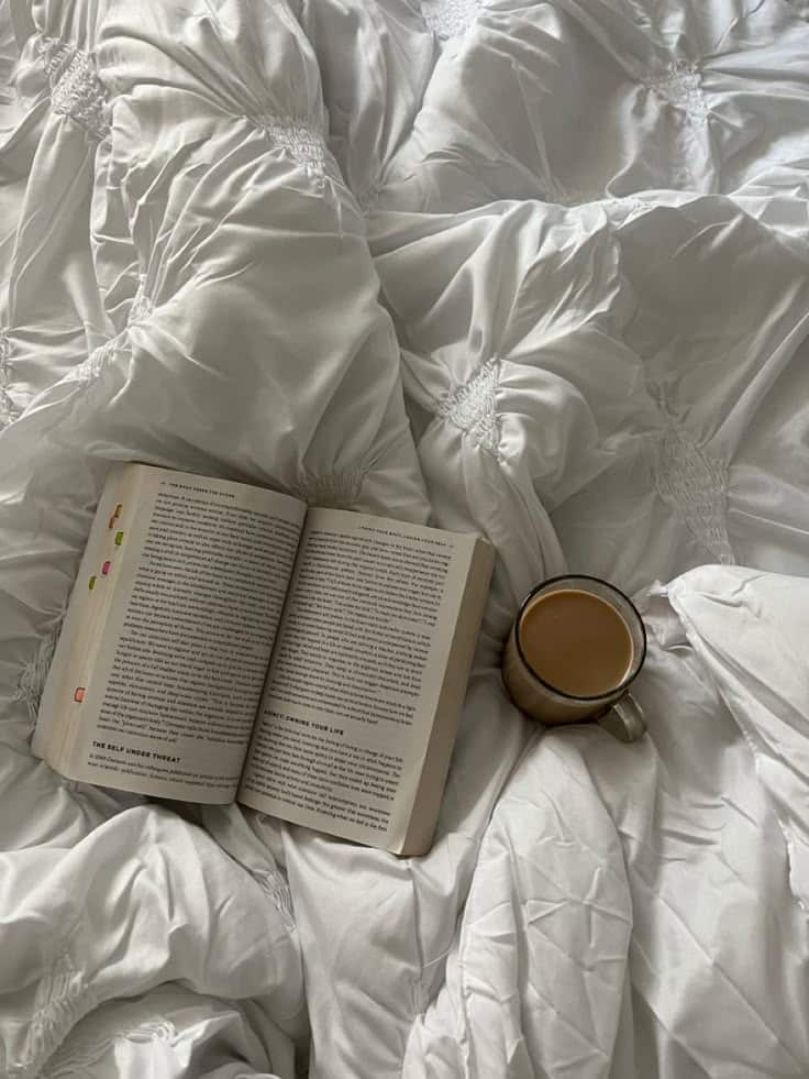 Cozy morning in bed with an open book and coffee on a white, textured comforter. Perfect relaxation scene.