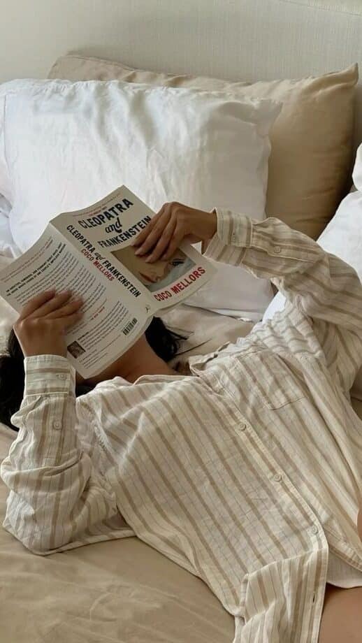 Woman reading a book in bed, wearing a striped shirt, surrounded by cozy pillows.