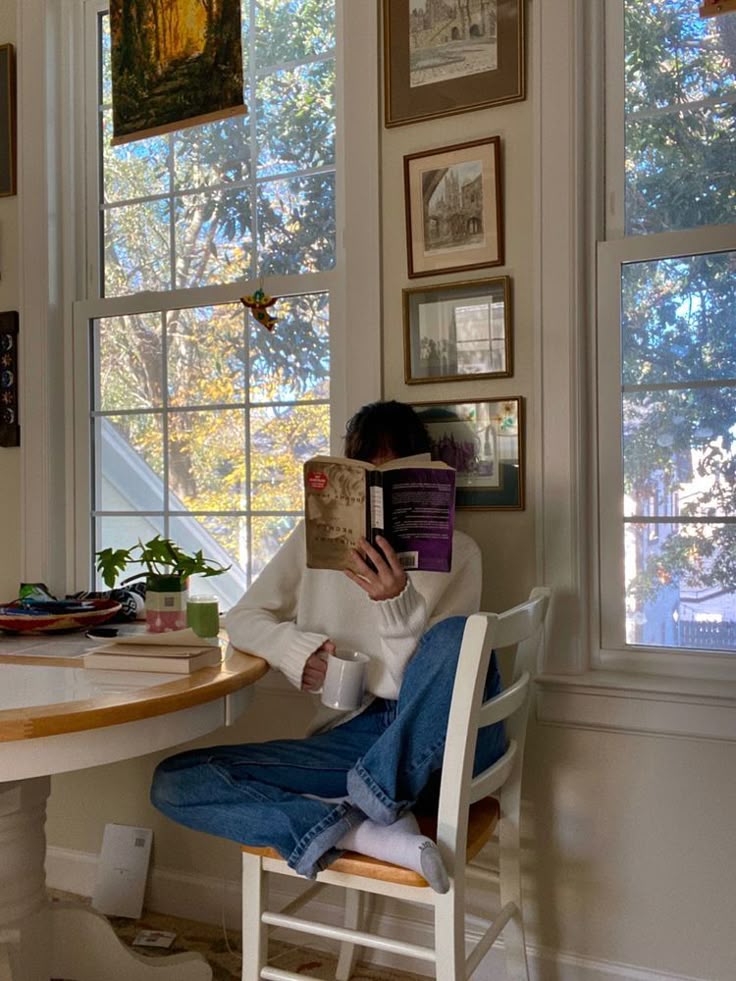 Cozy reading by a window with a hot drink, surrounded by sunlight and framed art. Perfect relaxation in a peaceful home setting.