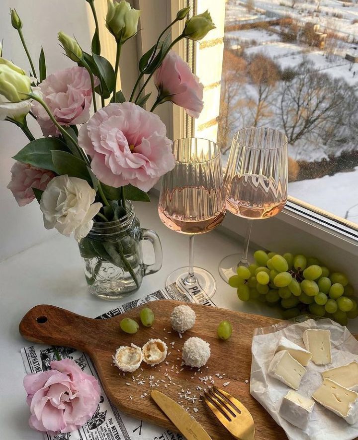 Elegant brunch scene: pink flowers, rosé wine, grapes, cheese, and sweets on a wooden board by a snowy window.