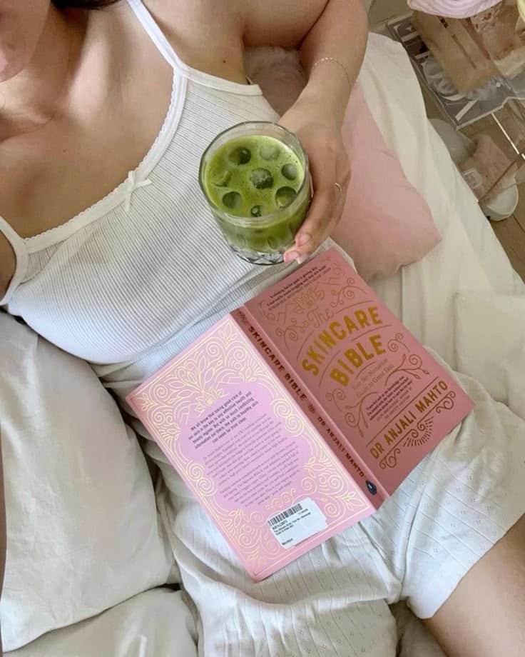 Woman relaxing with a skincare book and iced matcha drink in bed.