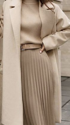 Elegant woman in a beige coat and pleated skirt, showcasing chic winter fashion against a neutral backdrop.