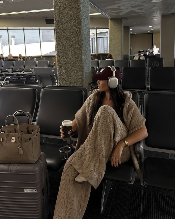 Person in cozy airport outfit with headphones, coffee, and luggage, relaxing in a terminal waiting area.