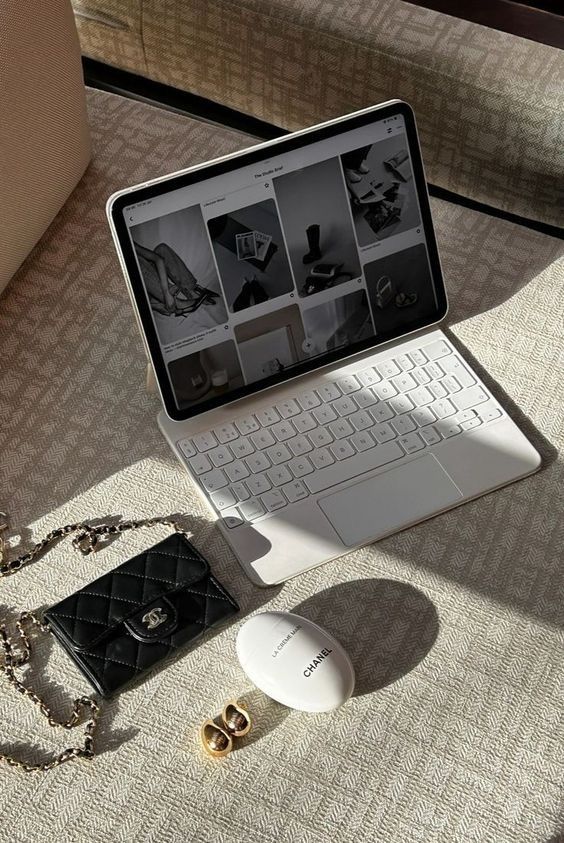 Tablet on beige textured surface with fashion accessories, including a black quilted wallet, Chanel hand cream, and small gold hoops.