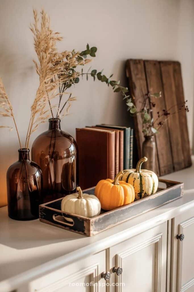 30+ Fall Decor Ideas That Make Any Room Feel Like Autumn - Handy Home Cozy fall decor with pumpkins on a tray, amber bottles, and autumn leaves on a white cabinet for seasonal home styling.