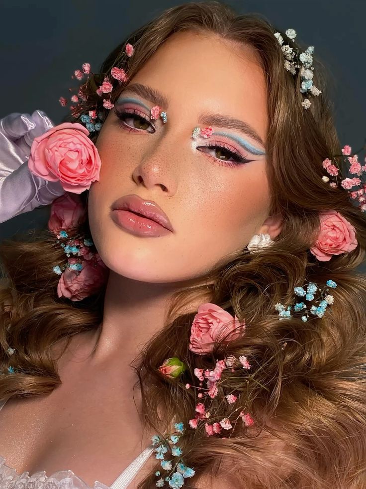 Young woman with colorful eye makeup, adorned with pink and blue flowers in hair, posed against a subtle background.