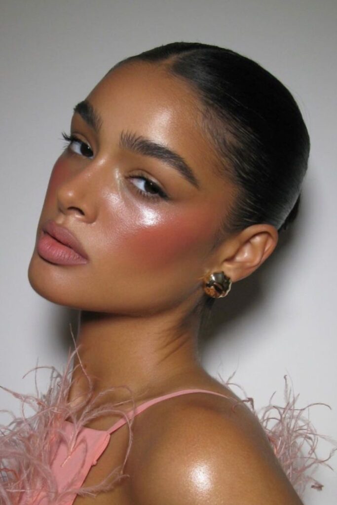 Glamorous woman with glossy makeup and sleek hairstyle, wearing a pink feathered top and stylish earrings.