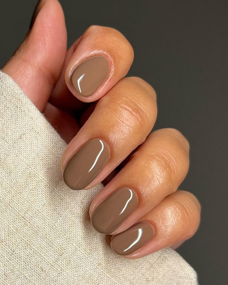 50e6dec5-136a-4b2c-b45e-785f2d130365 - Handy Home Close-up of a hand with glossy taupe nails on a beige fabric background, showcasing a modern manicure style.