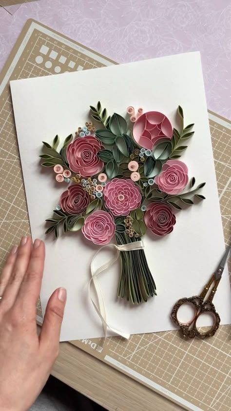 Quilled paper art bouquet with pink flowers and green leaves on a white background, featuring a hand and decorative scissors.
