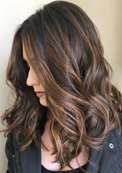 Woman with wavy, shoulder-length hair featuring caramel highlights, wearing a black jacket, looking down against a neutral background.