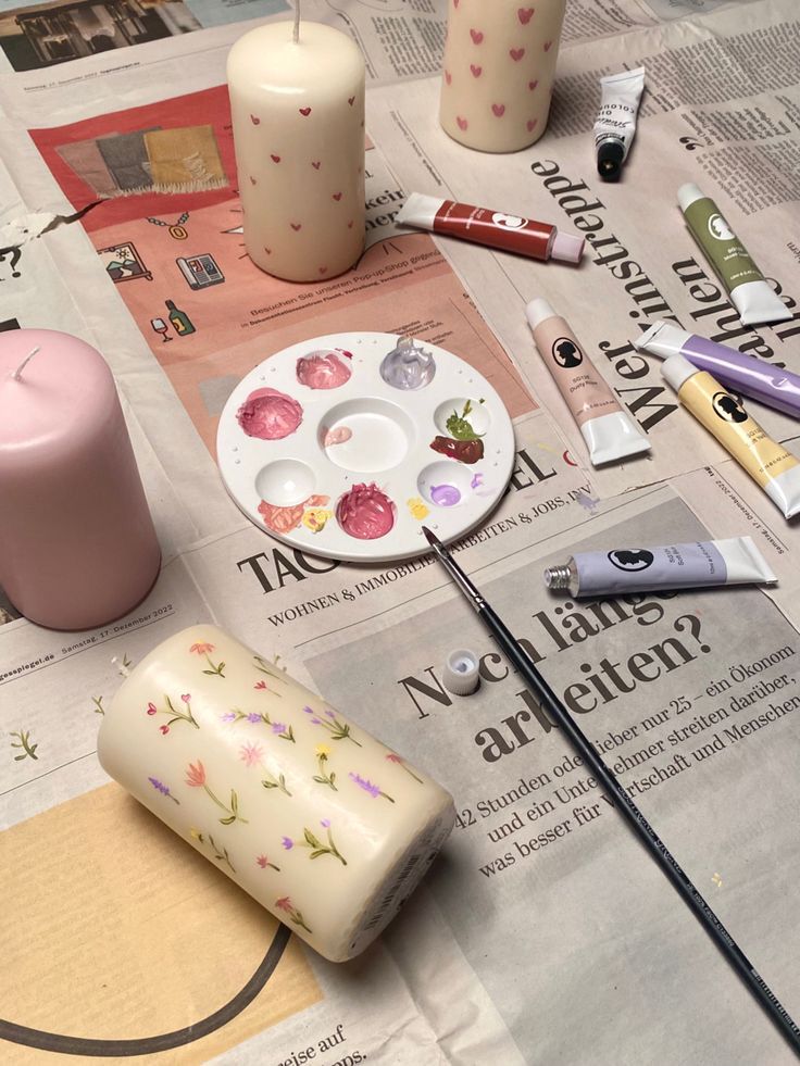 Candles being hand-painted with colorful designs on a newspaper-covered table, tubes of paint, and a paintbrush nearby.