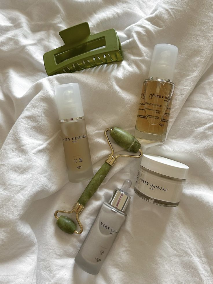 Skincare set with jade roller, cream, serum, hair clip on a white fabric. Luxurious beauty essentials flatlay.