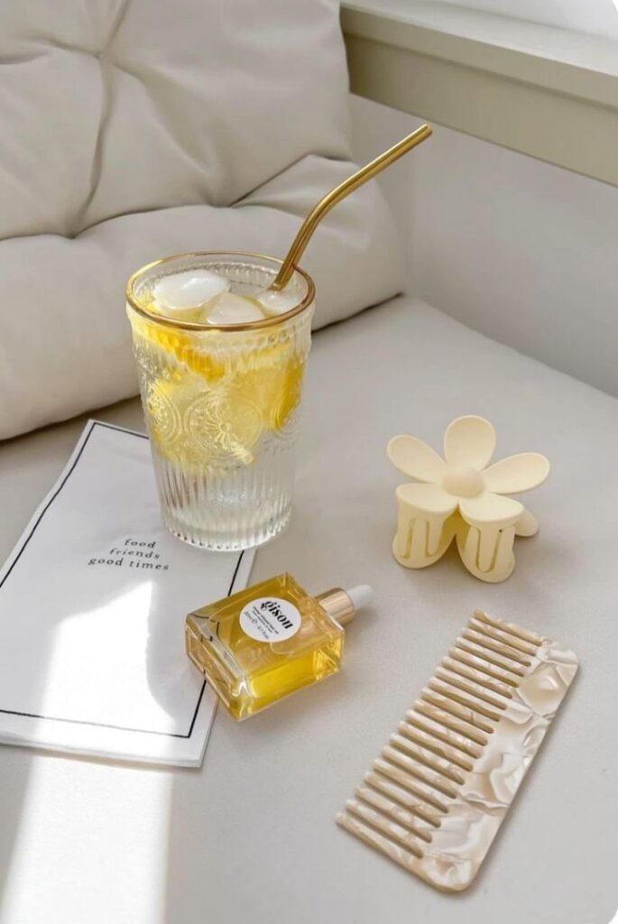 Glass of iced lemon water with a metal straw, hair oil, claw clip, and comb on a soft surface, sunlight streaming in. Relaxing vibes.