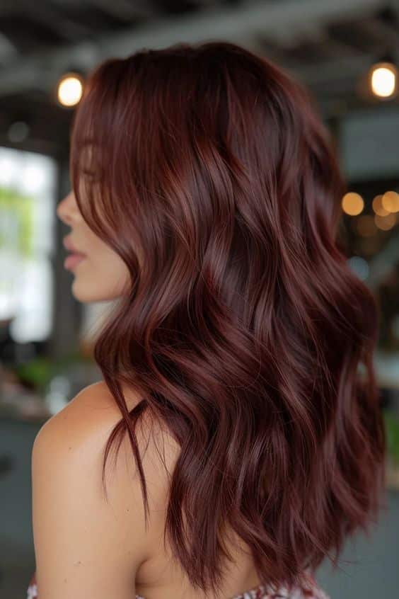 Woman with long, wavy burgundy hair in a stylish haircut, showcasing vibrant color and texture, in a bright salon setting.