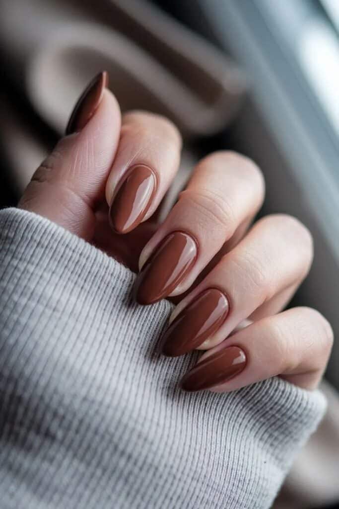 Close-up of a hand with sleek, brown manicured nails. Cozy gray sweater sleeve in a blurred background. Fashionable nail art style.