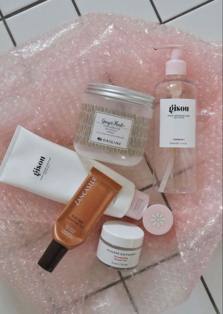 Assorted skincare and beauty products on pink bubble wrap, featuring Gisou hair wash and Origins Ginger Float cream.
