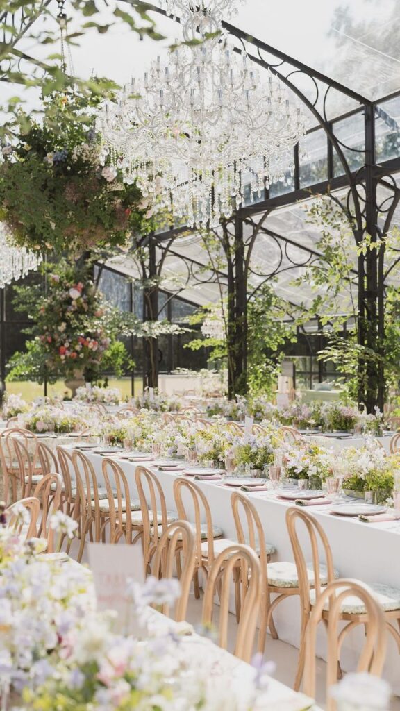 Elegant outdoor wedding reception with floral decor, crystal chandeliers, and wooden chairs under a glass canopy in a garden setting.