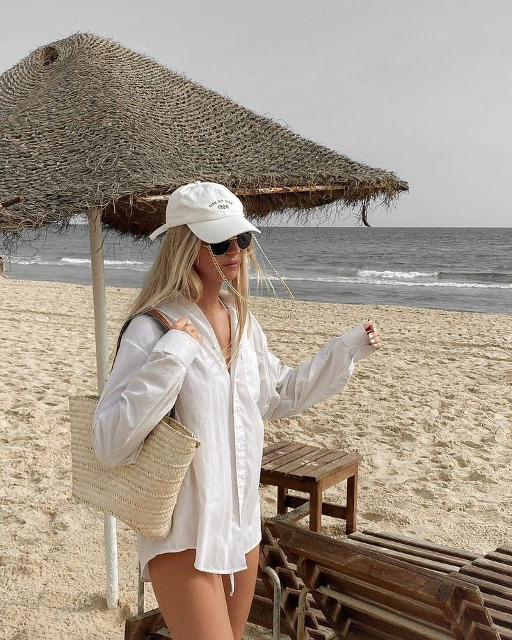 Woman in white shirt and cap with woven bag on a serene beach, under a straw parasol, with ocean waves in the background.