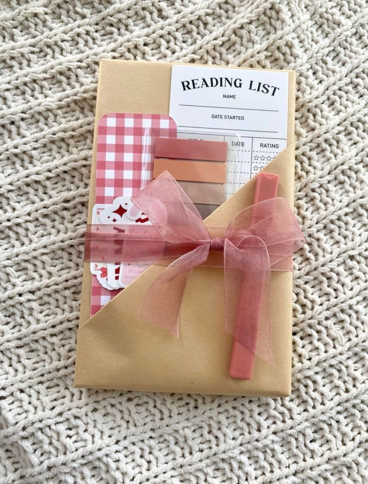 Gift package on a knitted blanket, featuring a reading list, pink marker, and decorative paper under a pink ribbon. Cozy reading essentials.