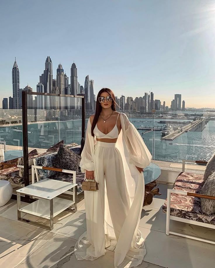 Fashionable woman in white outfit enjoying a sunny day on a Dubai rooftop with city skyline and marina view.