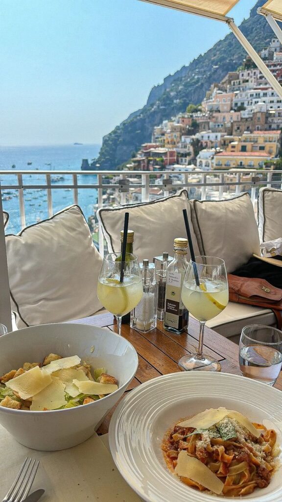 Elegant seaside meal with pasta and salad, overlooking a picturesque coastal village and the sparkling ocean in the background.