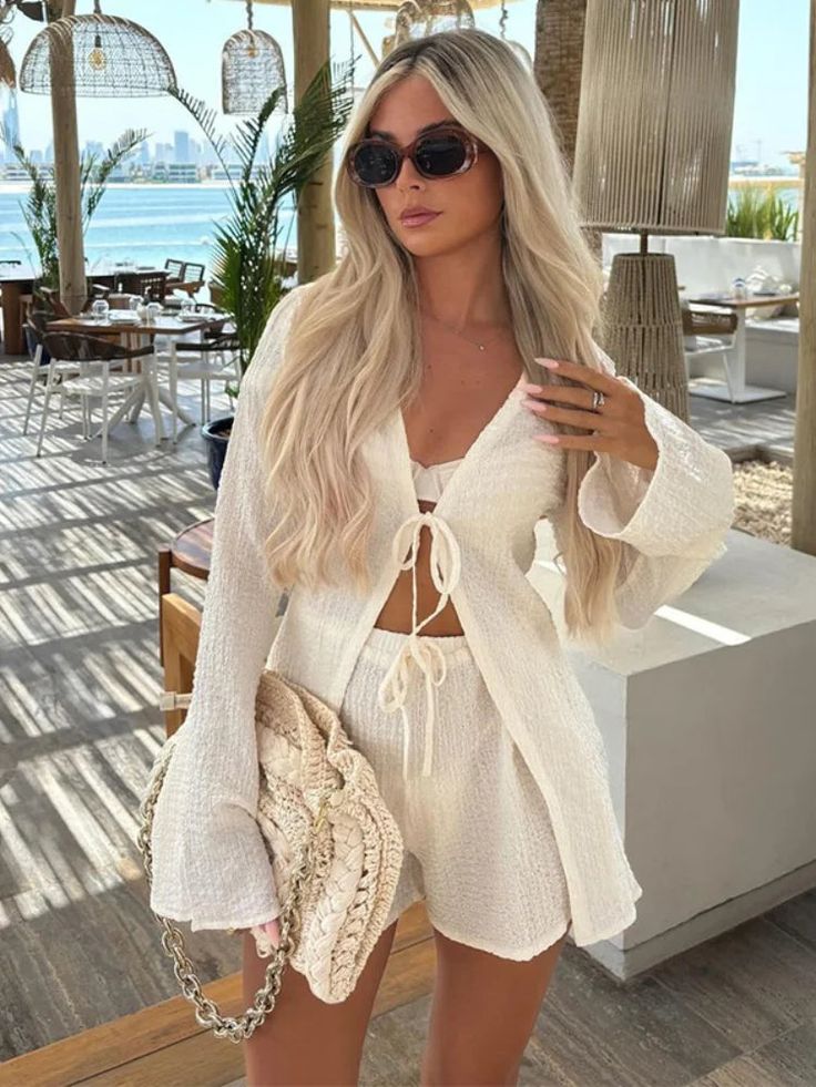 Blonde woman in chic white summer outfit with sunglasses and bag, sitting in an outdoor coastal cafe.