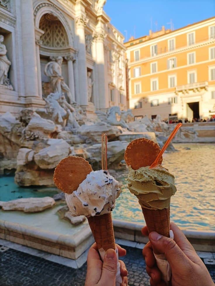 Two hands holding gelato cones with biscuits in front of the Trevi Fountain, Rome, basking in warm sunlight.