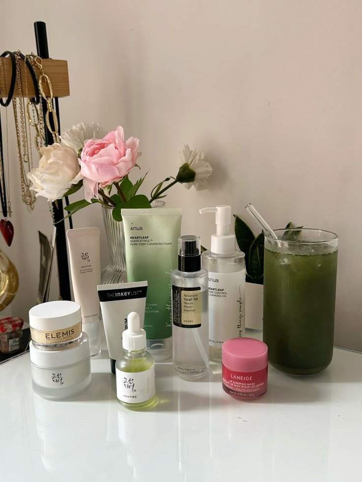 Skincare products on a white surface with flowers and a green drink, including serums, creams, and cleansing foam.