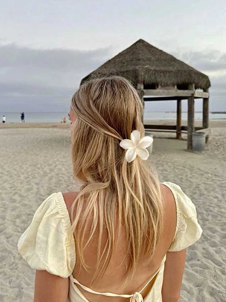 Woman in a yellow dress stands on a sandy beach, with a flower hair clip and a thatched hut in the background. Serene coastal scene.