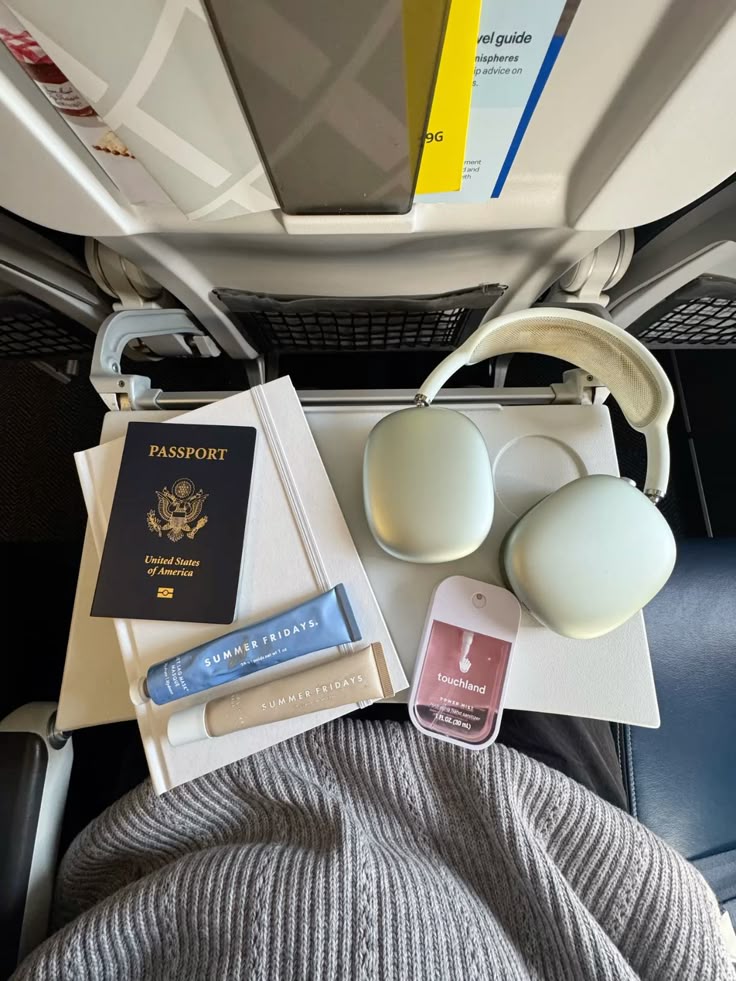 Travel essentials on plane tray: passport, headphones, skincare, hand sanitizer. Comfort and style for in-flight relaxation.