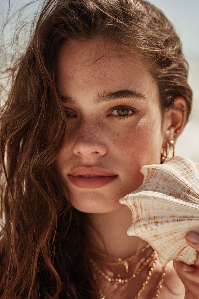Woman with freckles holding a shell by the beach, exuding natural beauty and summer vibes. Brown hair blowing in the wind.