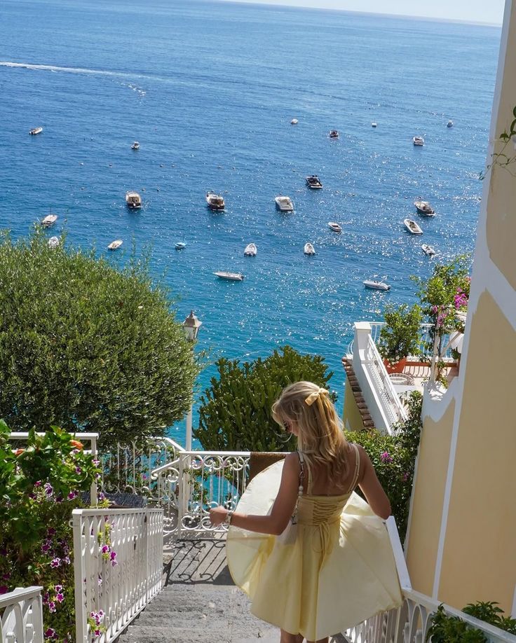 Woman in a yellow dress descending colorful Positano steps, overlooking sparkling sea with boats. Scenic Amalfi Coast vacation.