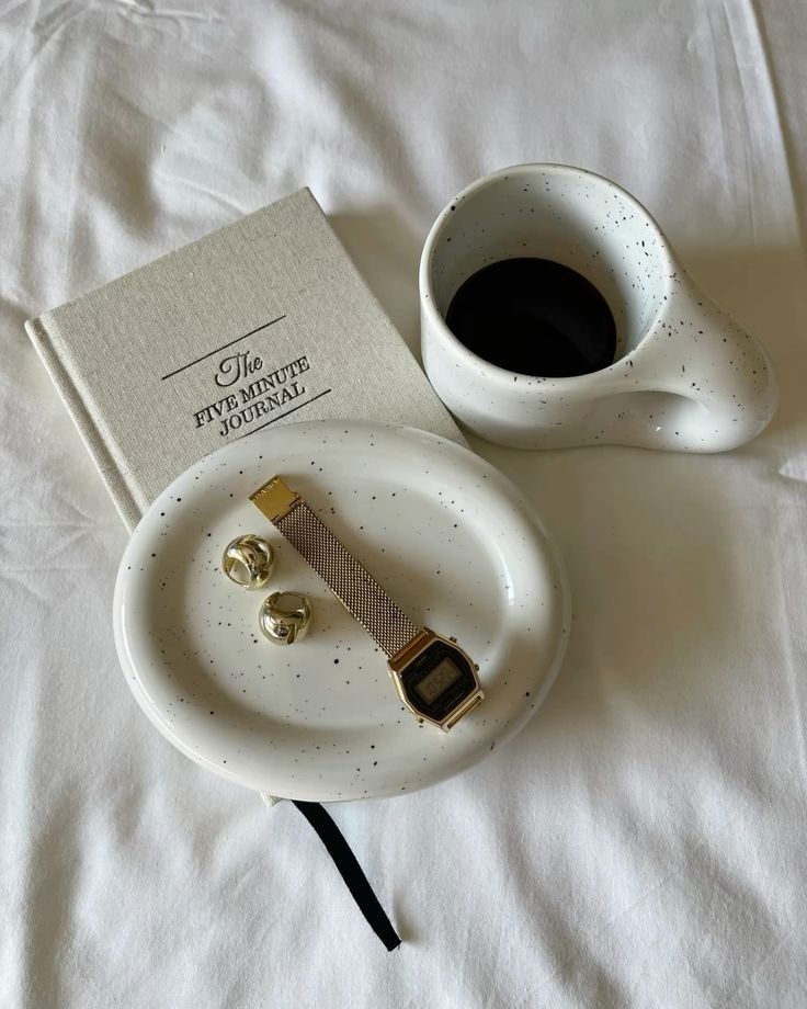 Minimalist setup with journal, coffee cup, golden watch, and rings on a tray, all on a white surface. Perfect for morning inspiration.