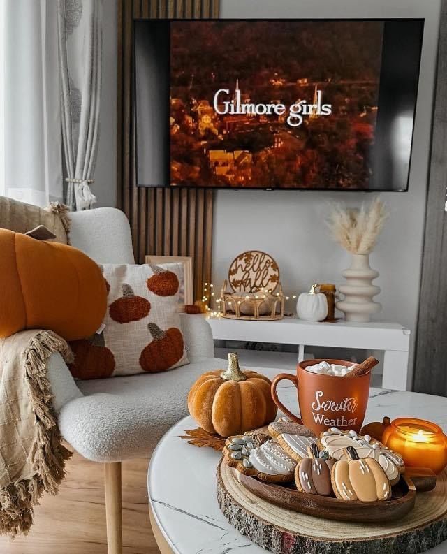 Cozy fall decor with pumpkin theme, Gilmore Girls on TV, pumpkin cookies, and hot cocoa creating a warm, autumn ambiance at home.