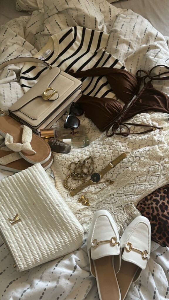 Flat lay of summer fashion essentials: striped shirt, swimwear, sandals, sunglasses, handbag, gold jewelry, and white loafers.
