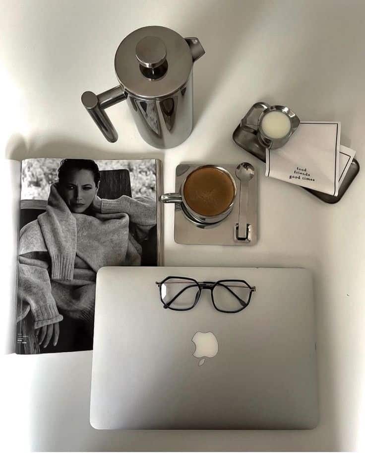 Stylish workspace with a laptop, glasses, coffee, magazine, and metallic French press on a white table. Cozy and modern setup.