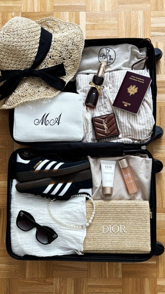 Packed suitcase with travel essentials: hat, sandals, passport, sunglasses, skincare, and accessories on wooden floor. Ideal for vacation.
