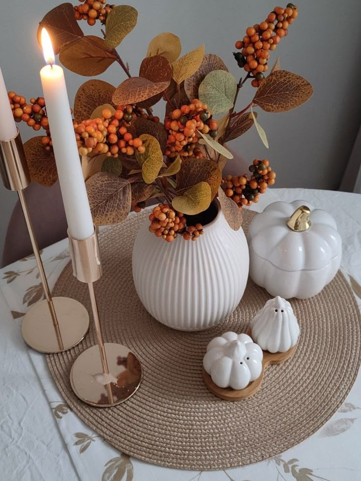 Elegant autumn table decor with candles, a white vase of orange berries, and ceramic pumpkins on a woven mat. Cozy fall ambiance.